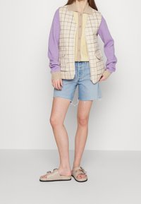 Beige och lila rutig väst med fickor över en gul topp, kombinerat med ljusblå denimshorts och beige glidsandaler med dekorativa spännen.