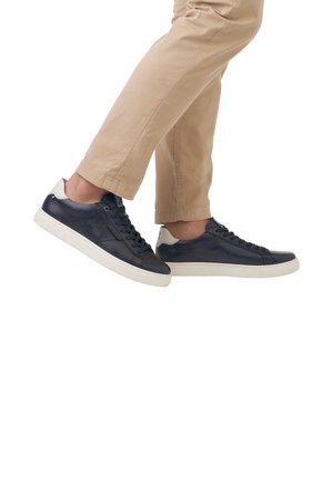 Zapatillas de cuero azul marino con suela de goma blanca, con un acabado suave y diseño de cordones, exhibidas en un pie con chinos beige.