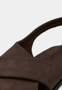 Καφέ σανδάλι από σουέντ με σχεδιασμό διασταυρούμενων λουριών, στρογγυλεμένες άκρες, μινιμαλιστική εμφάνιση και επίπεδη σόλα. Το υλικό με υφή φαίνεται απαλό.