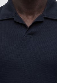 Κοντινό πλάνο του λαιμού και του πάνω μέρους του στήθους ενός ατόμου που φοράει μπλε σκούρο πόλο με υφή και εν μέρει ανοιχτό γιακά.