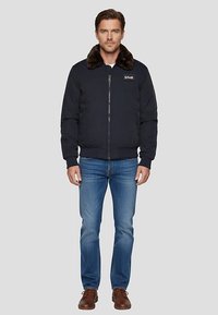 Giacca bomber blu navy con colletto in pelliccia, chiusura frontale con zip, polsini a costine e patch con logo sul petto. Indossata con jeans blu e scarpe marroni.