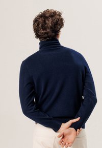 Marineblauer Rollkragenpullover aus weichem Strickmaterial, mit langenÄrmeln und taillierter Silhouette, getragen über hellen Hosen.