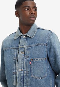 Jachetă din denim de culoare bleu deschis, cu închidere tip buton, guler ascuțit, buzunar pe piept și croială standard. Prezentă cusături în contrast și etichetă cu marcă.