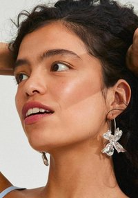 Silver butterfly-shaped earrings with a textured finish, featuring a hoop design. The model has dark, curly hair and a neutral background.