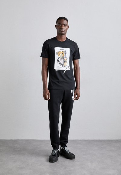 T-shirt noir à manches courtes avec un graphique d'ours aux accents colorés, associé à un pantalon de survêtement noir et des baskets noires.