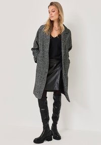 Szary, teksturowany długi płaszcz z otwartym przodem, zestawiony z czarną bluzką i skórzaną spódnicą. Czarne, wysokie botki do kolan dopełniają strój.