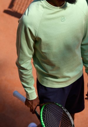 Personne portant un sweat-shirt vert clair et un short foncé tenant une raquette de tennis sur un court de tennis en terre battue.
