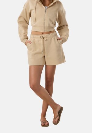 Donna che indossa felpa corta beige con zip e pantaloncini abbinati con coulisse, infradito marroni, in piedi con gambe incrociate e mani in tasca.
