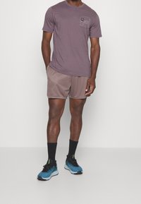 Hombre vistiendo una camiseta Under Armour color malva y pantalones cortos de malla, calcetines negros y zapatillas de correr azules, contra un fondo blanco liso.