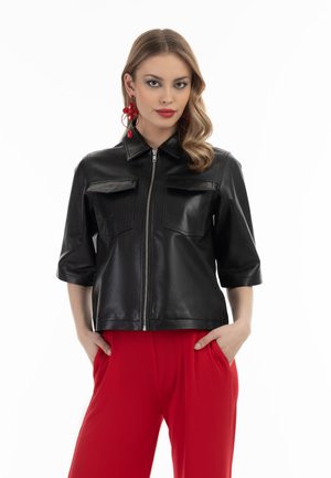 Giubbotto di pelle nera con zip frontale, maniche corte e due tasche sul petto. Indossato con pantaloni rossi, dal texture liscia.