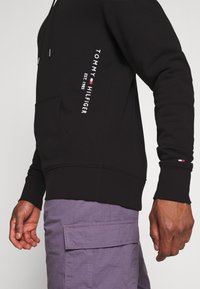 Sudadera negra con capucha y texto blanco "TOMMY HILFIGER" junto con detalle del logo en la manga. La tela parece suave y con un ajuste relajado.