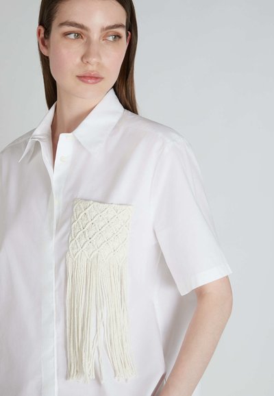 Camicia bianca a maniche corte con tasca in macramé bianco texturizzato con frange, chiusura a bottoni e design del colletto classico.