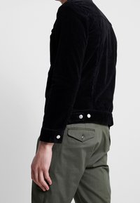 Svart kordflanelljacka med strukturerad yta, försedd med silverfärgade tryckknappar och en figursydd passform, kombinerad med olivgröna byxor.