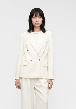 Jeune femme aux cheveux noirs et raides portant un blazer croisé écru avec des boutons dorés et un pantalon ample assorti.