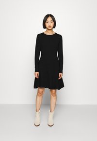 Vestido preto de mangas longas com corpete ajustado e saia rodada. Tecido suave, decote arredondado, usado com botas de tornozelo em creme.