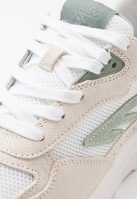 White and beige sneaker with mesh and suede materials. Features green accent, white laces, and textured details on the side.