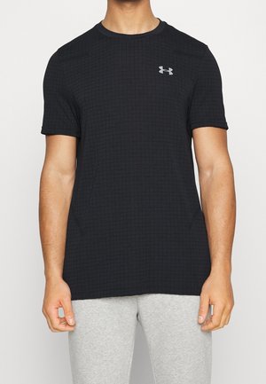 Mężczyzna ubrany w czarną, teksturowaną koszulkę z krótkim rękawem marki Under Armour oraz jasnoszare spodnie, stojący na tle gładkiej, białej ściany.