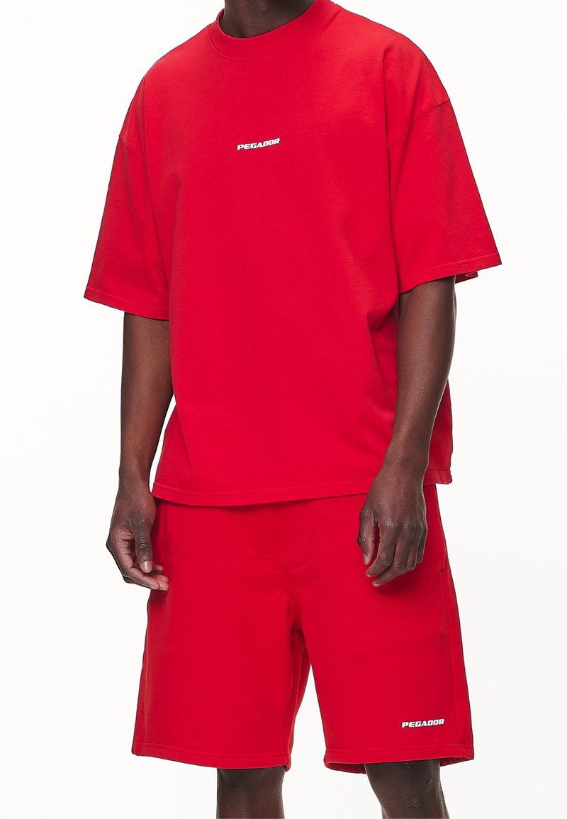 Ensemble t-shirt et short en coton rouge. Le t-shirt présente un col rond et des manches courtes, avec un logo blanc "PEGADOR" sur le devant.