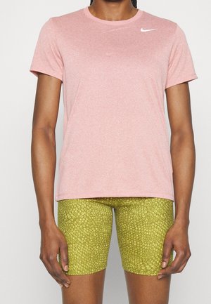 Personne portant un t-shirt Nike rose clair et un short de cycliste vert texturé, debout les bras détendus le long du corps.
