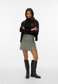 Un pull noir à col montant, une mini-jupe verte olive avec une fente sur le côté et des bottes en caoutchouc noires montantes. Les accessoires comprennent des chaussettes marron.