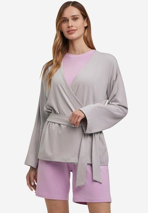 Vrouw draagt een lichtgrijze wikkelcardigan met een riem over een lavendelkleurig shirt en bijpassende lavendelkleurige shorts, staand tegen een witte achtergrond.