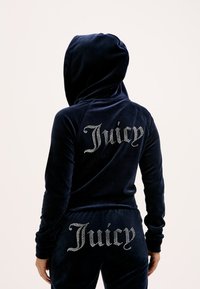Navyvelours trainingspak met capuchon. "Juicy" logo versierd met strass op de achterkant van het jack en de broek. Zachte textuur en aansluitend ontwerp.