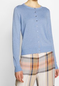 Frau in einer hellblauen Knopfjacke mit langen Ärmeln und karierten Hosen in Beige-, Orange-, Schwarz- und Blautönen.