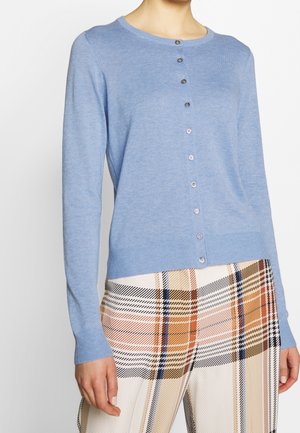 Kvinde iført en lys blå knap-åben cardigan med lange ærmer og rutede bukser i beige, orange, sort og blå nuancer.