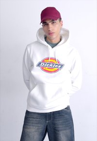 White hoodie with a colorful "Dickies" logo at the center, paired with blue jeans and a maroon cap. Hoodie features a front pocket.