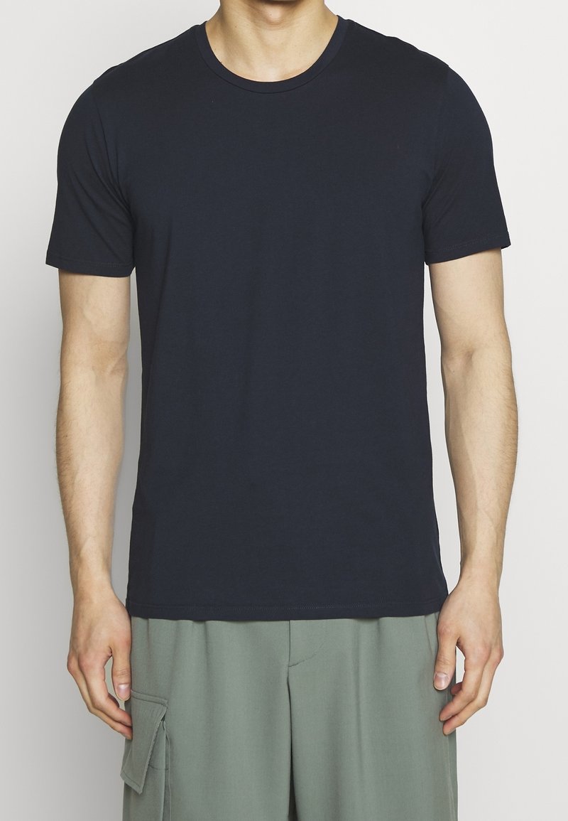 Mann, der ein einfaches dunkelblaues T-Shirt mit kurzen Ärmeln und hellgrüne Cargo-Shorts vor einem schlichten weißen Hintergrund trägt.