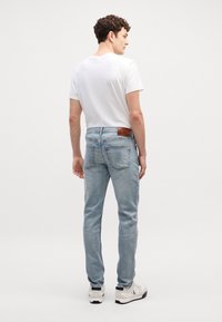 Jeans de mezclilla azul claro con un corte slim, que presentan un parche de cuero marrón en la parte posterior de la cintura y detalles de costura sutiles. Llevado con una camiseta blanca.