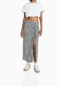 Floral Maxikleid in Schwarz und Weiß, mit seitlichem Schlitz und drapiertem Design. Kombiniert mit einem weißen Cropped-Top und Plateausneakern.