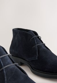 Marineblaue Wildleder-Stiefeletten mit rundlicher Zehenpartie, gesteppten Details und flachen schwarzen Gummisohlen. Mit Schnürverschluss und weicher Textur.