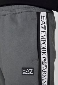 Grå sweatpants med svart midjeband, har en vit sidremsa tryckt med "EA7 EMPORIO ARMANI" och en liten logotypapplikation på benet.