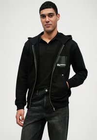 Sweatshirt preto com fecho, possui um bolso frontal, feito de material suave, combinado com uma camisa polo preta e jeans cinzentos, todos em um corte casual.