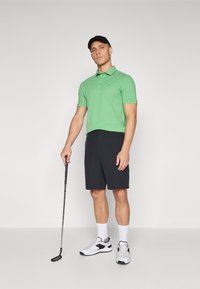 Ljusgrön polotröja med korta ärmar, svarta shorts, vita sportskor och en svart keps, hållande en golfklubba.