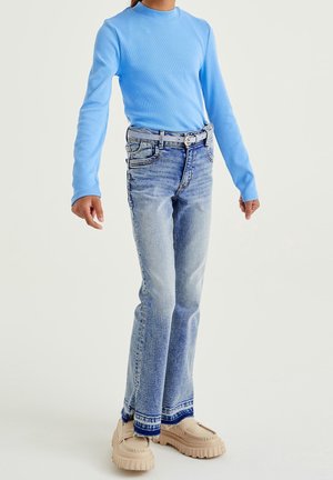 Osoba w jasnoniebieskiej koszuli z długim rękawem, wyblakłych niebieskich dżinsach z ciemnoniebieskimi wykończeniami, beżowych butach na masywnej podeszwie i szarym pasku.