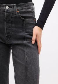 Χέρι που ακουμπάει στο επάνω μέρος του μηρού, φορώντας μαύρο εφαρμοστό τζιν και μακρυμάνικη μαύρη μπλούζα πάνω σε απλό φωτεινό φόντο.