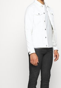 Chaqueta de denim blanca con dos bolsillos en el pecho, botones de presión plateados y un color clásico, llevada sobre una camisa gris y jeans oscuros.