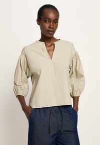 Frau trägt eine beige V-Ausschnitt Bluse mit lockeren, gepufften 3/4 Ärmeln und dunkelblauben Jeans mit Kordelzug, die Hände in den Taschen, neutraler Hintergrund.