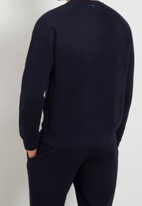 Man som bär en mörk marinblå sweatshirt och matchande byxor, står med ena handen i fickan, ryggen mot kameran.