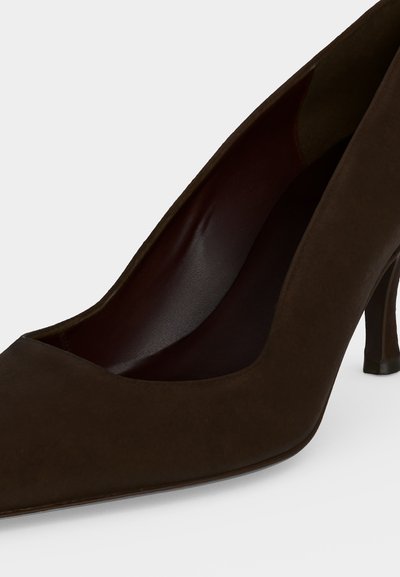 Chaussure à talon haut pointu en daim marron foncé avec un bord supérieur incurvé et un talon aiguille fin sur fond blanc.