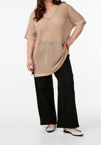 Beige gebreide tuniek met een V-hals en korte mouwen, met een losse pasvorm en een getextureerd patroon, gecombineerd met zwarte wijde broekspijpen en beige schoenen.