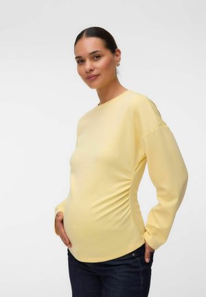Femme enceinte portant un haut jaune à manches longues et un jean foncé, tenant doucement son ventre arrondi et souriant à la caméra.