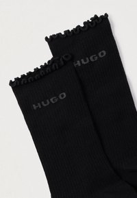 Čierne rebrované ponožky s volánovým lemom; na každej ponožke je sivé logo s nápisom "HUGO". Mäkký, elastický materiál.
