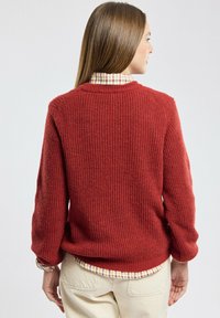 Pull en tricot rouge avec une texture côtelée, un col rond et des épaules tombantes, porté par-dessus une chemise à carreaux avec un ourlet arrondi.