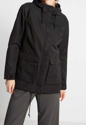 Person wearing a black hooded jacket with front pockets and drawstrings, paired with loose gray pants, standing against a plain background.