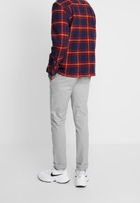 Pantaloni grigi a sigaretta con una texture liscia, abbinati a una camicia flanellata a scacchi nei colori blu navy, rosso e arancione, indossati con sneakers bianche.