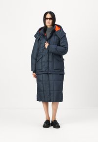 Cappotto imbottito blu navy con cappuccio arancione, dotato di tasche frontali e gonna lunga fino al ginocchio. Tessuto testurizzato con design trapuntato.