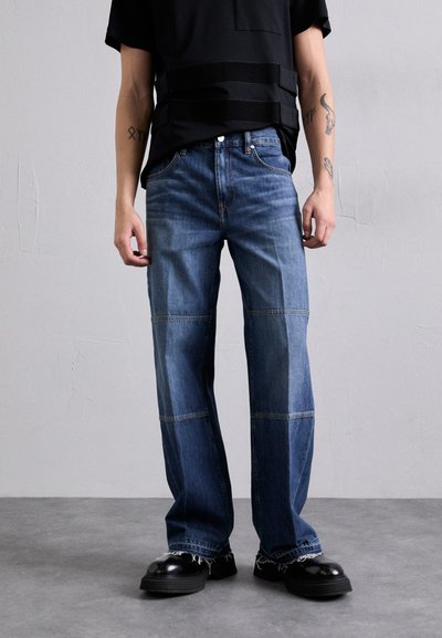 Jean en denim bleu avec une coupe droite et des coutures visibles. Présente des panneaux contrastants et des ourlets effilochés. Porté avec des chaussures noires.